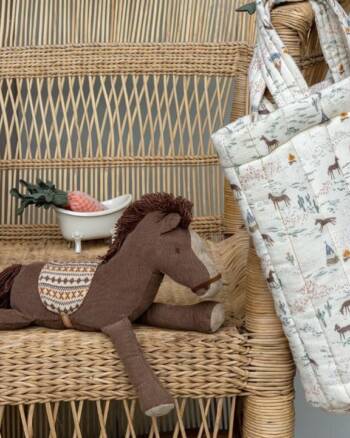 Doudou Poney Maileg allongé sur un fauteuil en rotin, à côté d’un sac imprimé enfant