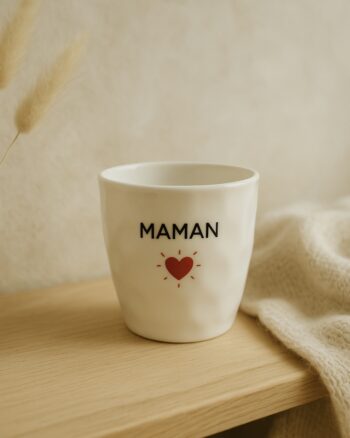 Gobelet en céramique blanche avec le mot MAMAN et un cœur rouge entouré de petits traits, posé sur une table bois clair