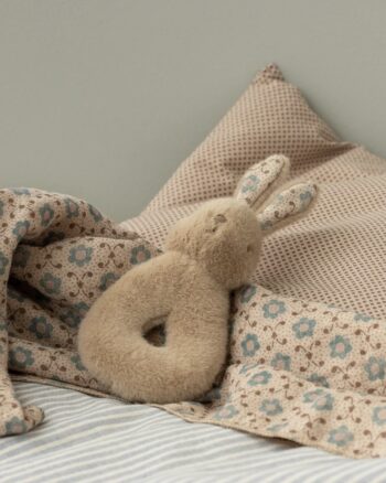 Hochet lapin Maileg crème pêche posé sur un lit avec une couverture imprimée fleurie