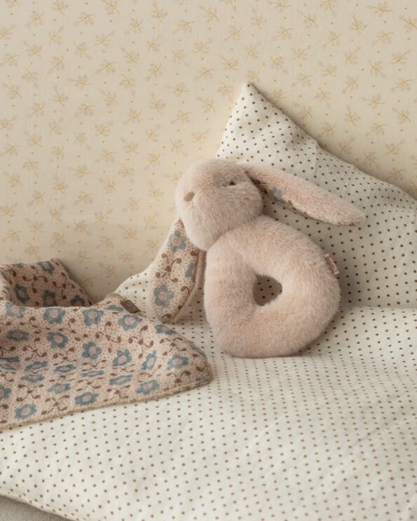 Hochet lapin Maileg rose poudre couché sur un lit avec lange imprimé et coussin à pois
