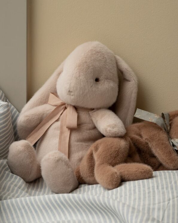 Peluche Bunny Maileg Small rose poudré posée sur un lit à rayures avec peluche marron