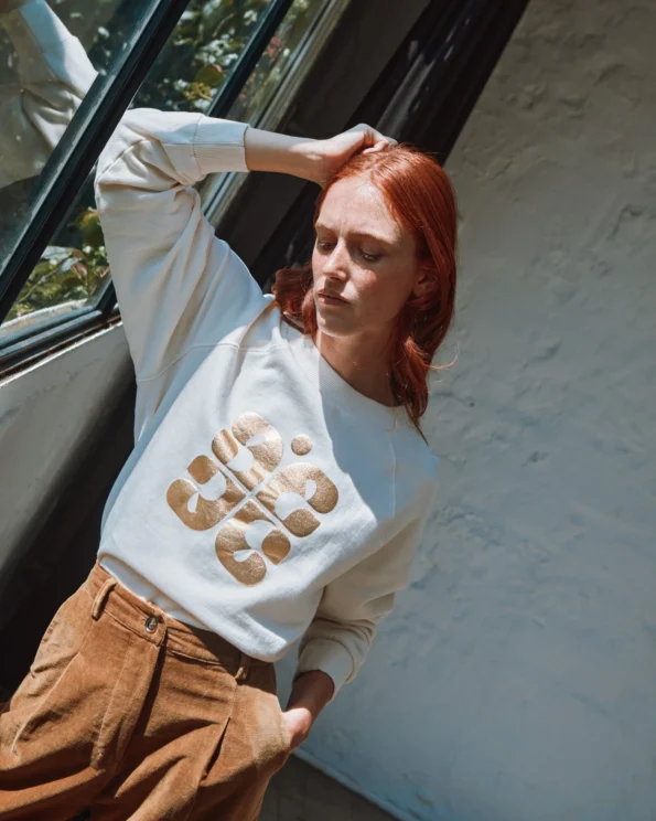 Sweat crème logo doré Emile et Ida – porté par une femme près d’une fenêtre