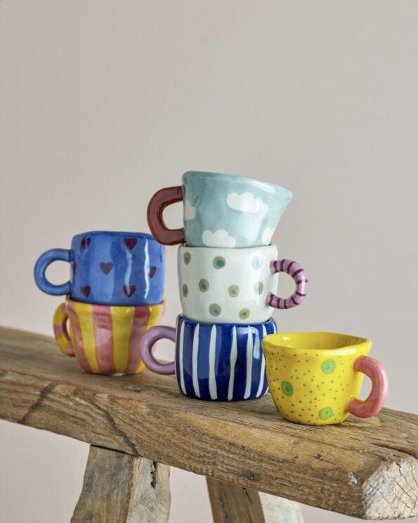 Collection de tasses Nini Bloomingville en grès peint à la main, empilées sur un banc en bois