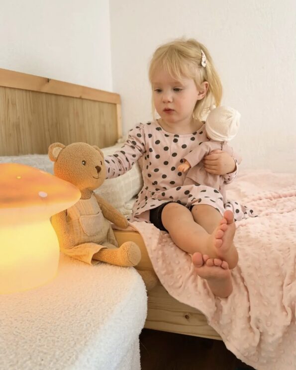 Enfant jouant avec la peluche Marcel Ours Egmont Toys sur un lit, accompagnée d’une veilleuse champignon