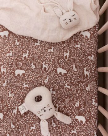 Hochet Lapin Écru Main Sauvage tricoté à la main, posé dans un lit bébé sur drap brun à motifs