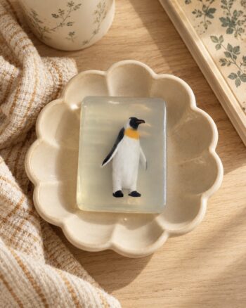 Savon pingouin Ludi Soap transparent présenté dans une coupelle fleur en céramique beige.