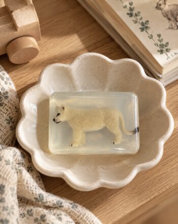 Savon lionceau beige Ludi Soap transparent dans une coupelle fleur en céramique beige sur fond de bois naturel.